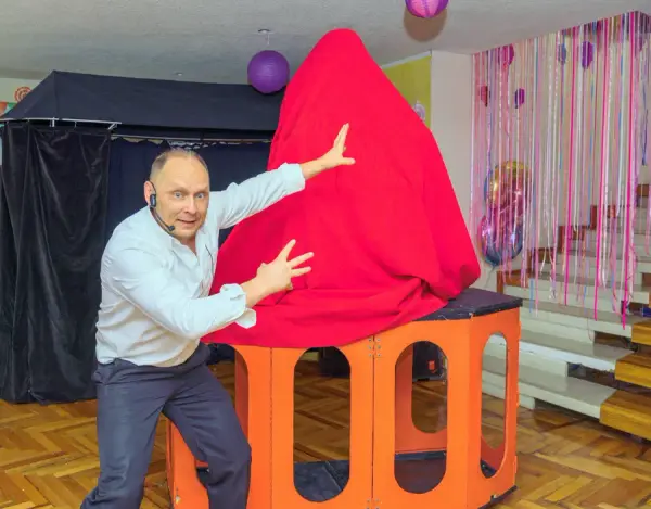 El mago señala con gesto dramático una gran ilusión cubierta de tela roja